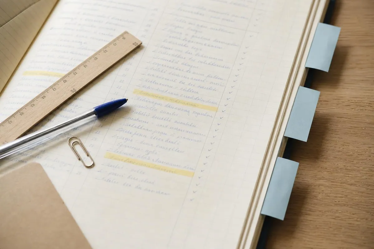 Gros plan sur un carnet à colonnes ouvert où des dates sont surlignées en jaune paille, avec des onglets adhésifs bleus, une règle plate et un stylo posés en travers